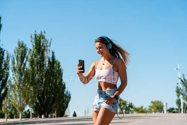 Patinadora feminina positiva em patins e fones de ouvido tomando auto-tiro no telefone celular no dia ensolarado no verão na cidade — Fotografia de Stock