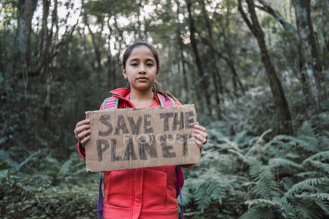Ethnic child showing Save The Planet title on carton piece while looking at camera in forest — Stock Photo