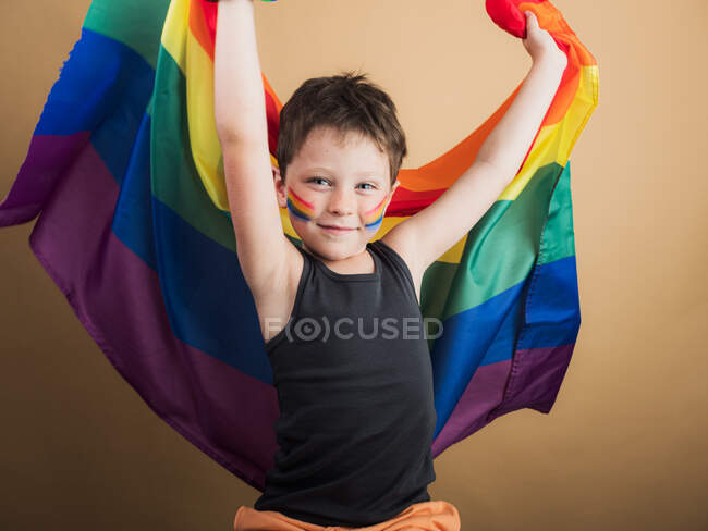 Веселый ребенок с макияжем на щеках, поднимающий флаг ЛГБТК, глядя в камеру на бежевом фоне — стоковое фото