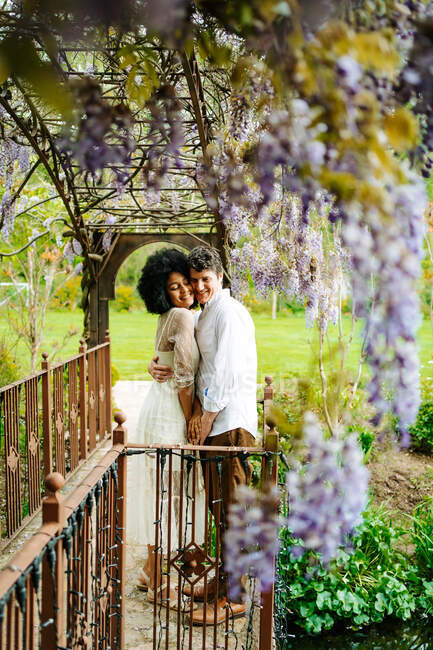 Вид збоку ніжної багаторасової пари, що обіймається, стоячи з очима, закритими під аркою з квітучими фіолетовими квітами вістерії в саду — стокове фото