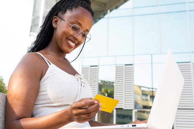 Low angle of cheerful African American female making payment with plastic card while using laptop during online shopping in Barcelona street — Stock Photo