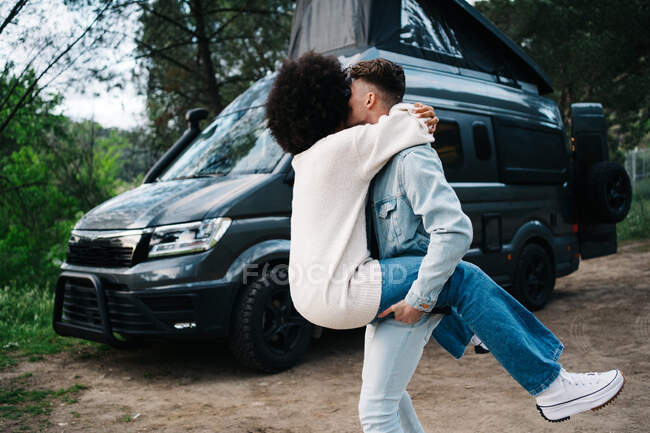 Cheerful young African American woman laughing happily and embracing boyfriend while having fun together near camper van parked in green forest during summer journey together — Stock Photo