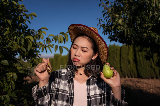 Sad ethnic female farmer standing with ripe pear bitten by insects in garden in village — Stock Photo