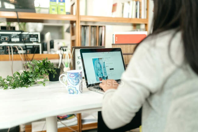 Back view of female using netbook during online shopping while sitting at table with hot drink at home and choosing goods — Stock Photo