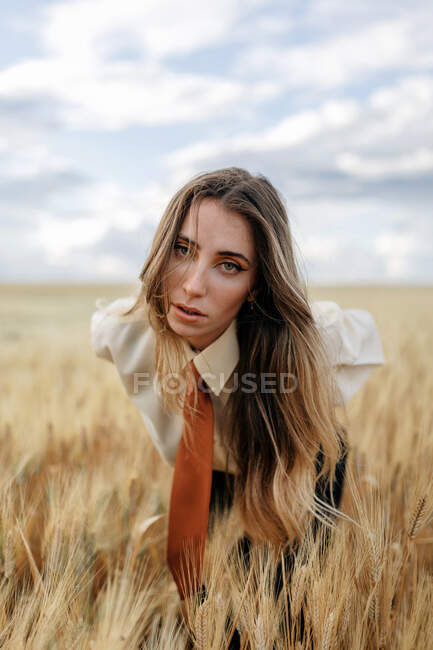 Joven hembra con cabello ondulado mirando a la cámara inclinándose hacia adelante en el campo bajo el cielo nublado sobre un fondo borroso - foto de stock