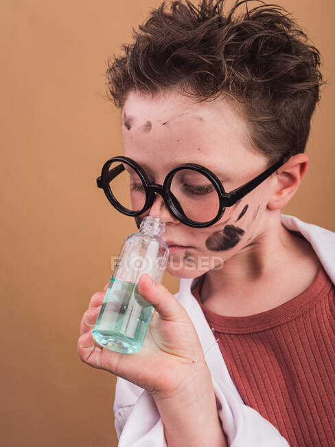 Enfant chimiste dans des verres en plastique avec des taches de peinture sur le visage sentant le liquide de la bouteille sur fond beige — Photo de stock