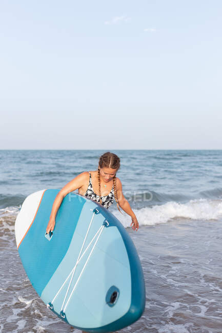 Mujer en traje de baño de pie con tabla SUP en agua de mar en verano y mirando hacia abajo - foto de stock