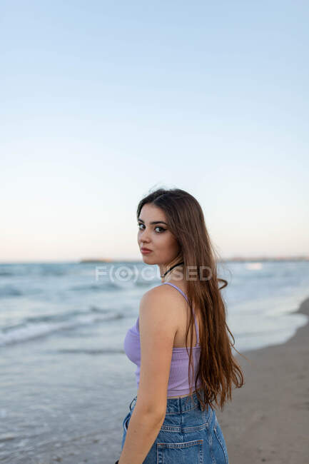 Мечтательная молодая женщина с длинными волосами смотрит в камеру, стоя на песчаном пляже рядом с морем. — стоковое фото
