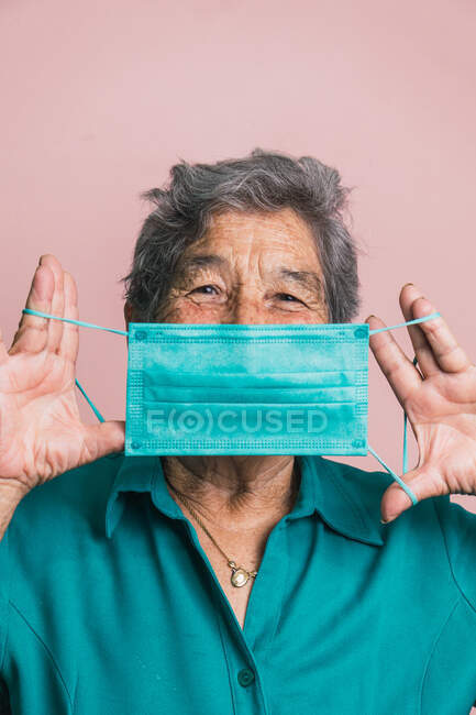 Smiling aged female covering mouth with blue protective medical mask from coronavirus while looking at camera on pink background in studio — Stock Photo