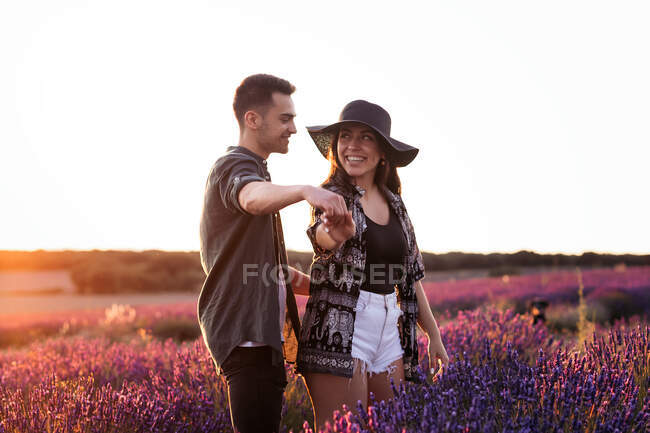 Vista lateral del hombre sosteniendo la mano de su novia en sombrero mientras se miran entre sí contra el florecimiento de lavanda en la espalda iluminada - foto de stock