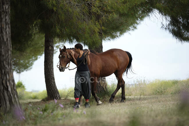 Full body of cheerful ethnic female stroking obedient brown stallion with bridle while walking together on green meadow with trees in summer countryside — Stock Photo