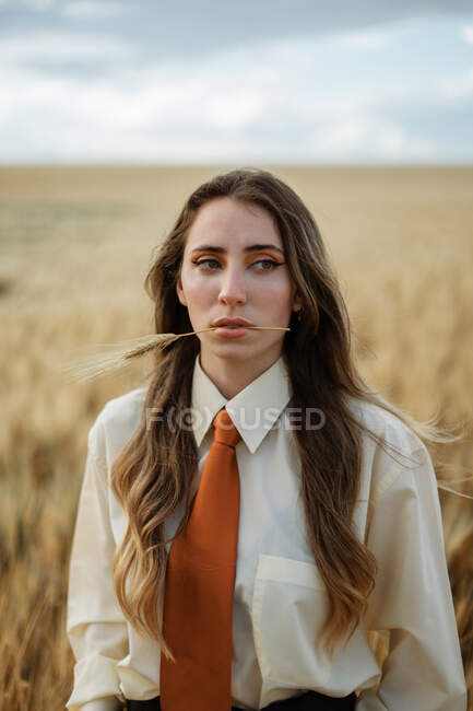 Jeune femme contemplative dans des vêtements élégants avec cravate et pointe dans la bouche regardant loin dans la campagne — Photo de stock
