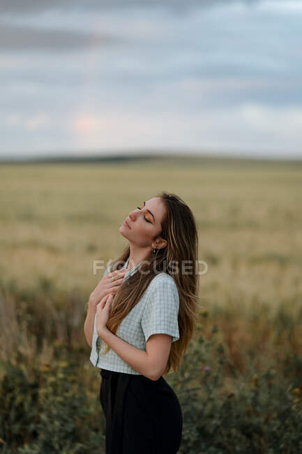 Вид збоку на молоду усвідомлену жінку з закритими очима на дорозі біля лугу під хмарним небом ввечері в сільській місцевості — стокове фото