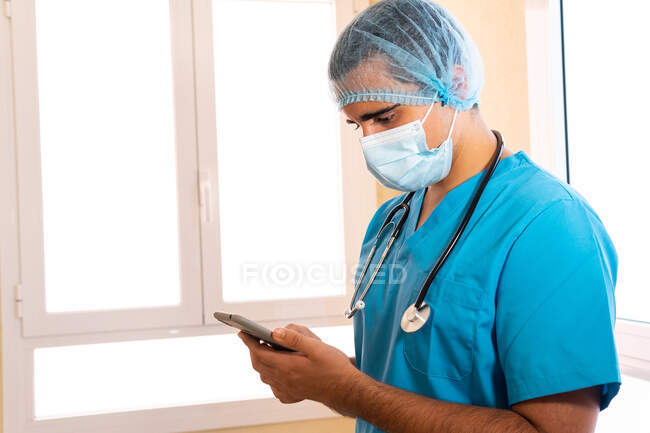 Male medic in uniform and with stethoscope standing in hospital and surfing Internet on smartphone — Stock Photo