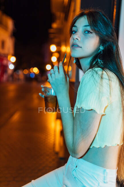 Content young female with long hair and glass of refreshing drink looking at camera at dusk — Stock Photo
