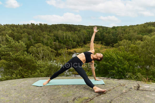 Все тело босиком женщина делает Патита Тарасана позировать на мат, практикуя йогу на камне в природе — стоковое фото