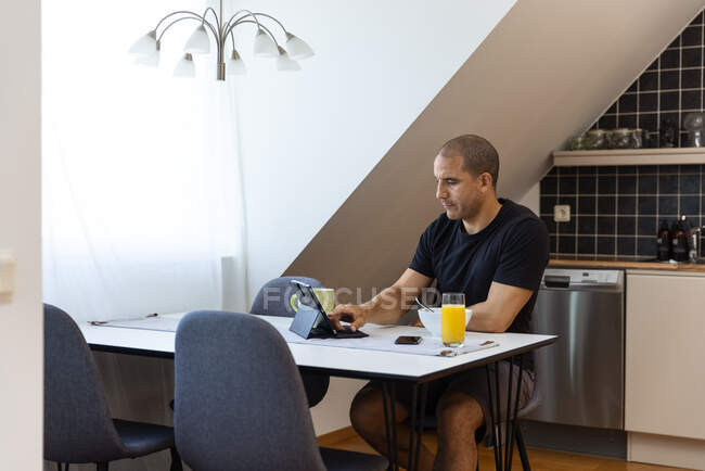 Focused male surfing Internet on tablet while sitting at table at home and enjoying breakfast in morning — Stock Photo