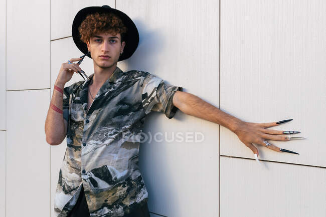 Hombre joven vano en ropa elegante con sombrero de pie en la pared de baldosas mirando a la cámara - foto de stock
