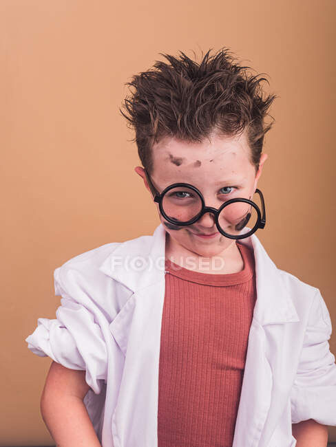 Child with dirty face and uncombed hair in decorative glasses looking at camera on beige background — Stock Photo
