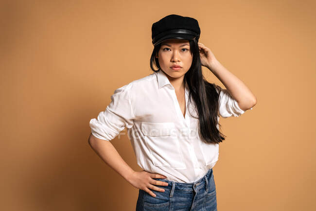 Вид спереди на модную азиатскую модель в белой рубашке и джинсах, касающихся кепки на бежевом фоне и смотрящую в камеру — стоковое фото