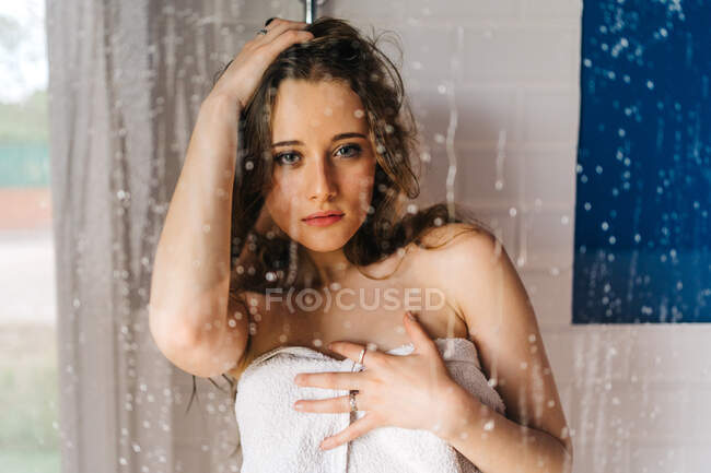 Weibchen in weißes weiches Handtuch gehüllt, steht hinter nasser Glastür der Duschkabine und blickt in die Kamera — Stockfoto