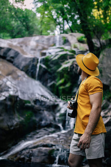 Vue latérale d'un randonneur masculin méconnaissable debout sur un rocher et admirant une cascade dans une forêt — Photo de stock