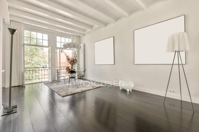 Moderno loft diseño interior de la casa con mesa de comedor y sillas colocadas cerca de la ventana en la esquina de la habitación espaciosa con imágenes simuladas colgando de la pared blanca en la casa - foto de stock
