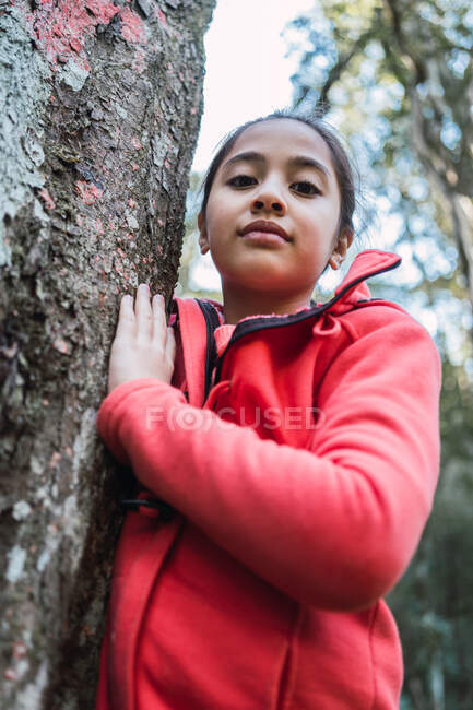 From below of charming ethnic child touching rough bark of aged tree trunk with lichen while looking at camera in forest — Stock Photo