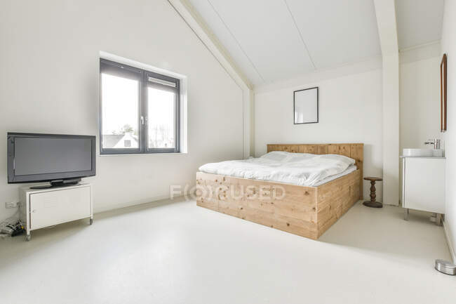Luz ático dormitorio interior con paredes blancas amuebladas con cama con TV en esquina en casa de estilo loft moderno - foto de stock
