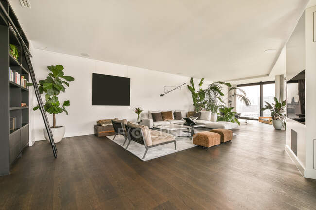 Дизайн интерьера стильной просторной гостиной с большим телевизором, висящим на белой стене перед столом и мягкими диванами, украшенными растениями в современной роскошной квартире — стоковое фото