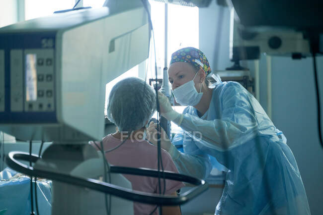 Mujer atenta cirujana en uniforme estéril examinando ojo de paciente anónima contra refractómetro en el hospital - foto de stock