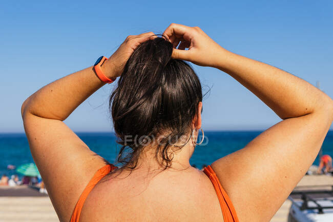 Back view of anonymous plump female athlete in sportswear making a ponytail against ocean under blue sky — Stock Photo
