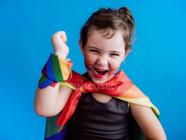 Милый счастливый ребенок с разноцветной повязкой на шее и запястье стоя на синем фоне и глядя на камеру — стоковое фото