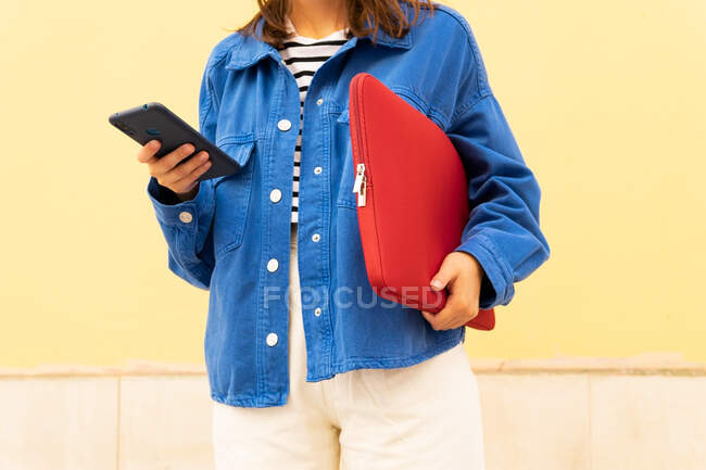 Crop female student with netbook in case browsing mobile phone while standing against yellow wall in city street — Stock Photo