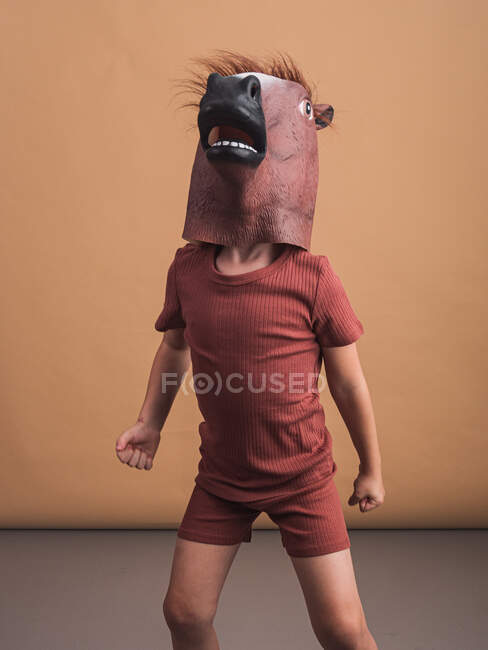 Niño anónimo en máscara de caballo que representa el concepto de semental galopante sobre fondo beige - foto de stock
