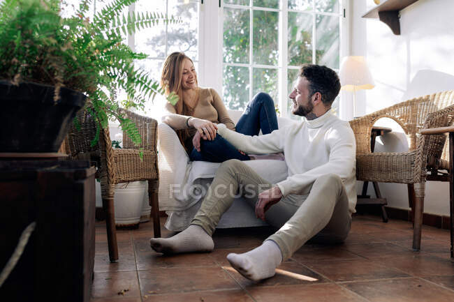 Homem barbudo no chão e conteúdo namorada em poltrona falando enquanto olha um para o outro em casa — Fotografia de Stock