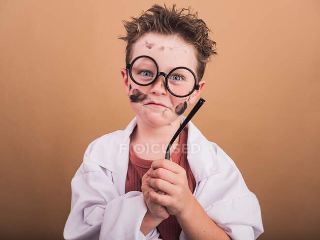 Niño con manchas de pintura en la cara y anteojos templo en bata de laboratorio mirando a la cámara sobre fondo beige - foto de stock