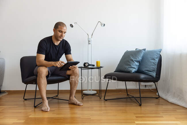 Hombre adulto enfocado sentado en sillón cerca de la mesa con un vaso de jugo de naranja y el uso de tabletas en la mañana en casa - foto de stock