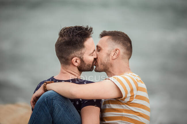 Adult bearded man in striped t shirt embracing and kissing homosexual boyfriend with closed eyes on blurred background — Stock Photo