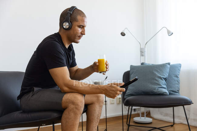 Side view from above of male in headphones watching video on tablet while drinking orange juice and sitting on chair in morning at home — Stock Photo