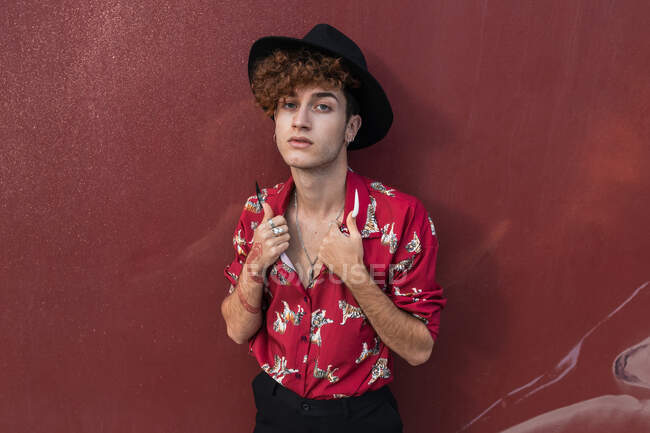 Jeune homme homosexuel élégant en chemise ornementale et chapeau avec de longs ongles et tatouage regardant la caméra — Photo de stock
