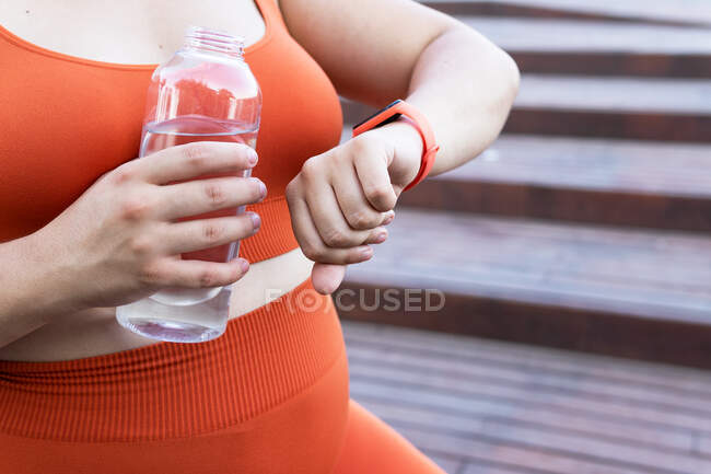Crop unrecognizable plus size female athlete with bottle of aqua watching heart rate on wearable tracker during workout on urban stairs — Stock Photo