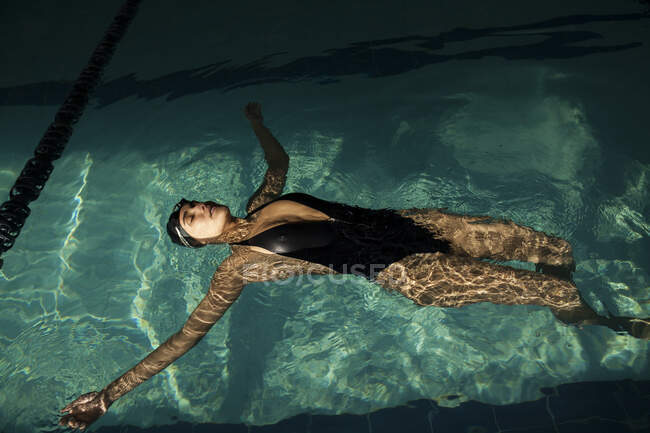 Jovem mulher bonita dentro da piscina interior, vestindo maiô preto, flutuando de costas — Fotografia de Stock