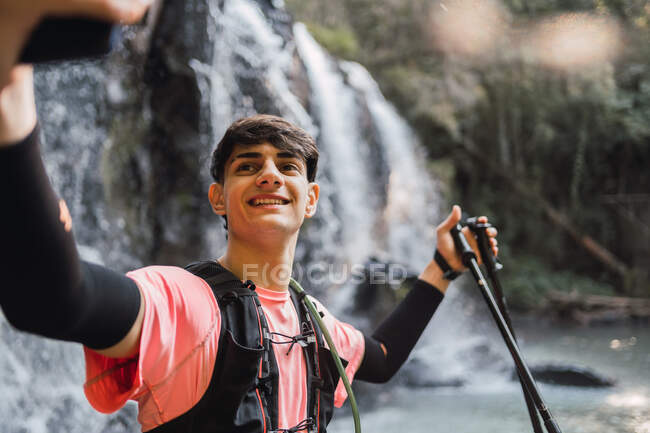 Усміхнений чоловічий пішохід робить самостріл на смартфоні, стоячи на тлі водоспаду та озера в лісі під час походів — стокове фото