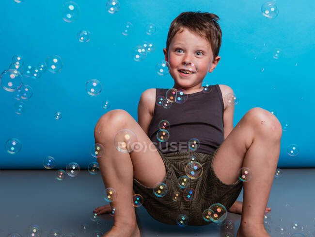 Surpreendido pré-adolescente criança sentado no chão e olhando para bolhas de sabão voador em estúdio em fundo azul — Fotografia de Stock