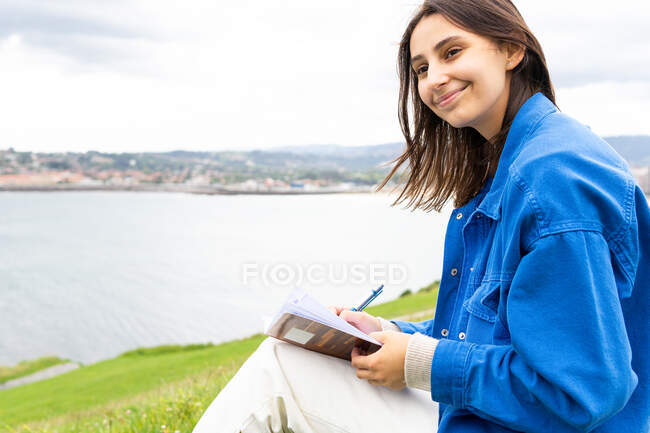 Happy female writing in notepad while sitting on hill near sea and looking away — Stock Photo