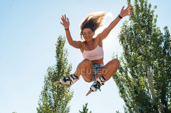 De abaixo da fêmea ativa em patins saltando e realizando truque no parque contra o céu azul no verão no dia ensolarado — Fotografia de Stock
