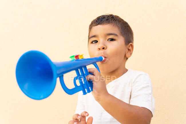 Netter kleiner Junge spielt blaue Spielzeugtrompete, während er in die Kamera schaut und in der Nähe einer hellorangen Wand steht — Stockfoto