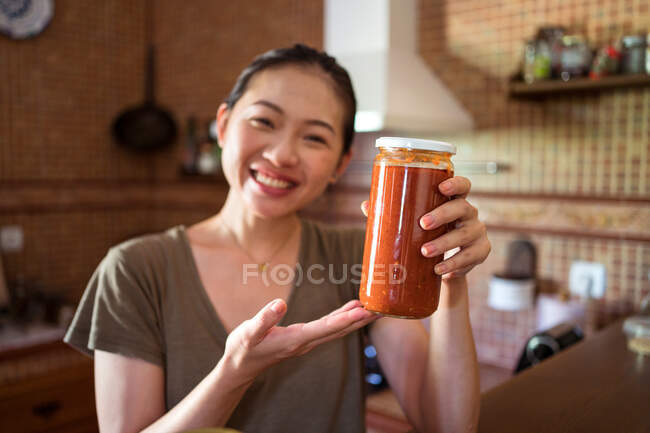 Fröhliche ethnische Hausfrau zeigt Glasgefäße mit hausgemachter Tomaten-Marinara-Sauce, während sie am Tisch in der Küche sitzt und in die Kamera blickt — Stockfoto
