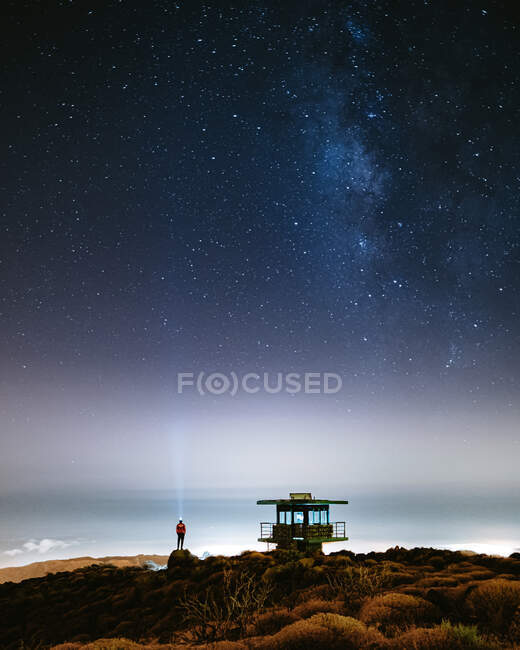 Distant view of person standing on seashore near rescue tower under spectacular night sky with glowing stars of Milky Way — Stock Photo